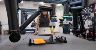 A technician with long dark hair standing at a robot work cell with a laptop, under black ceiling-mounted robot arms, with orange parts in a fixture on the bench and an American flag in the background