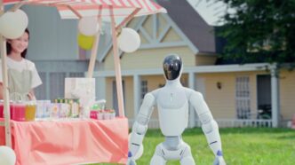 White Galbot ET1 humanoid robot with a glossy black visor standing on a lawn beside a pink-striped drinks stand with balloons, a smiling girl behind the stall