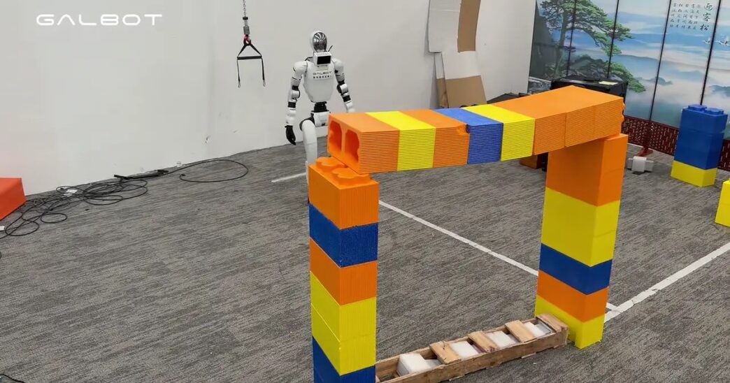 White Galbot humanoid robot stands behind an arch of oversized orange, yellow and blue blocks in a lab with a GALBOT watermark