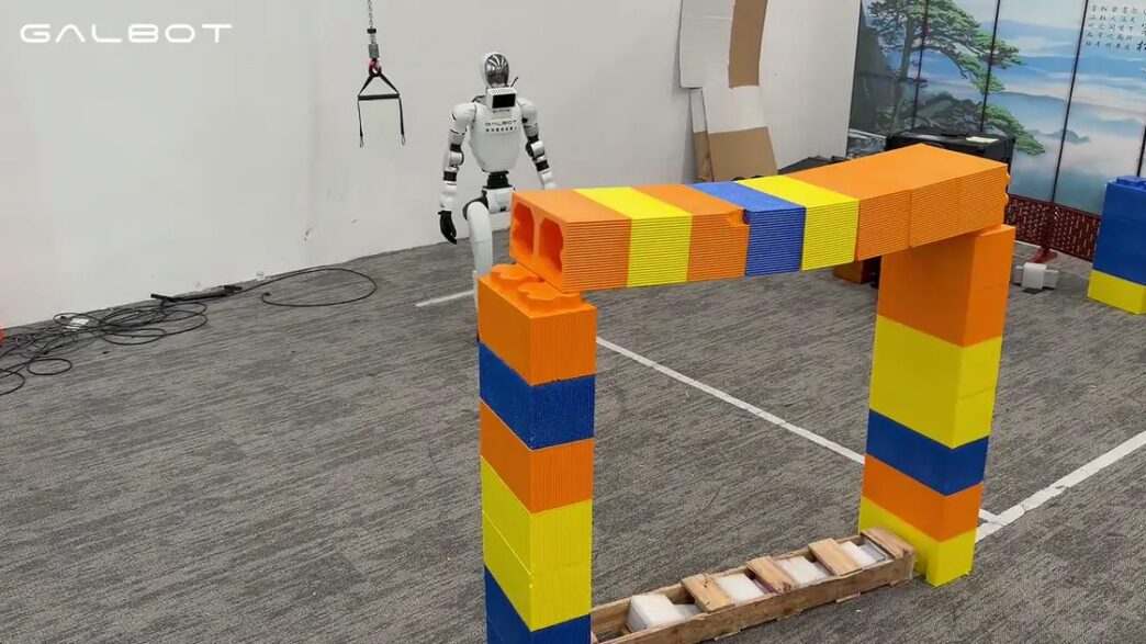 White Galbot humanoid robot stands behind an arch of oversized orange, yellow and blue blocks in a lab with a GALBOT watermark