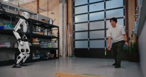 Apptronik Apollo humanoid robot in a lab throws toward a wooden cornhole board while a person in a white shirt watches, with overhead support cables visible