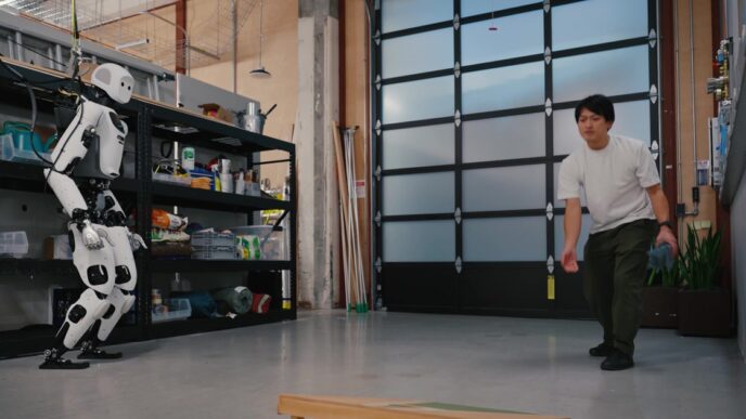 Apptronik Apollo humanoid robot in a lab throws toward a wooden cornhole board while a person in a white shirt watches, with overhead support cables visible