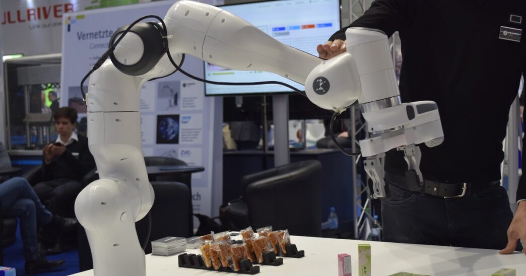 White Franka Emika collaborative robot arm with a parallel gripper over snack packages on a trade-show table