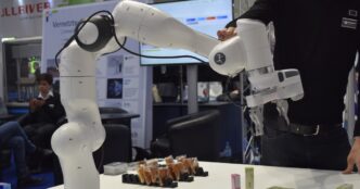 White Franka Emika collaborative robot arm with a parallel gripper over snack packages on a trade-show table