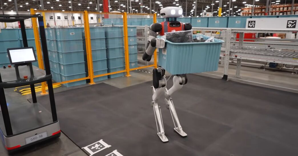 Agility Robotics' Digit humanoid robot, with a white head and red chest, walking across a warehouse floor carrying a blue plastic tote, past orange safety fencing, stacked totes and a conveyor