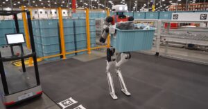 Agility Robotics' Digit humanoid robot, with a white head and red chest, walking across a warehouse floor carrying a blue plastic tote, past orange safety fencing, stacked totes and a conveyor
