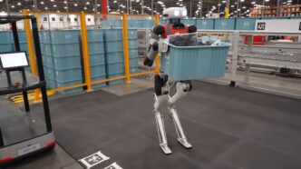Agility Robotics' Digit humanoid robot, with a white head and red chest, walking across a warehouse floor carrying a blue plastic tote, past orange safety fencing, stacked totes and a conveyor