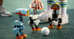 Three small Microduck robots, one light blue, one white and one dark grey, with orange feet standing around a speckled ball on a green mesh floor while a person in tall striped socks reaches in