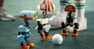 Three small Microduck robots, one light blue, one white and one dark grey, with orange feet standing around a speckled ball on a green mesh floor while a person in tall striped socks reaches in