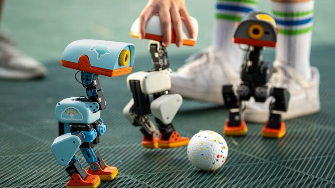 Three small Microduck robots, one light blue, one white and one dark grey, with orange feet standing around a speckled ball on a green mesh floor while a person in tall striped socks reaches in
