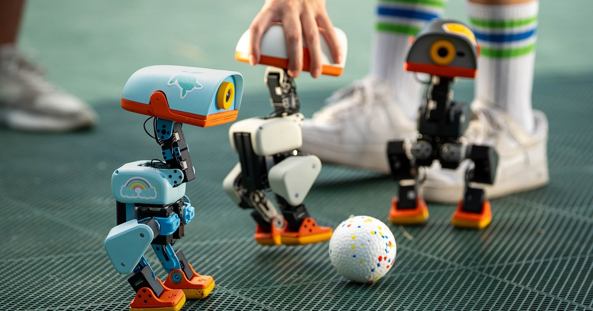 Three small Microduck robots, one light blue, one white and one dark grey, with orange feet standing around a speckled ball on a green mesh floor while a person in tall striped socks reaches in