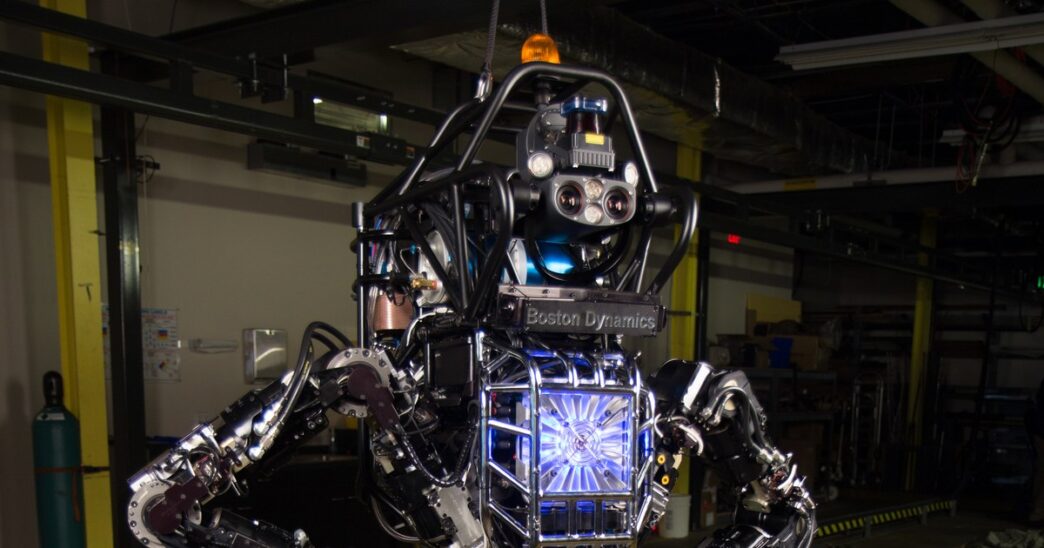 Hydraulic Boston Dynamics Atlas humanoid from the DARPA program in a test bay, exposed hydraulic joints, hoses and sensor head visible