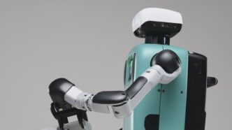 Close view of an Agility Robotics Digit humanoid reaching forward with one jointed arm, teal torso and white head unit against a grey backdrop