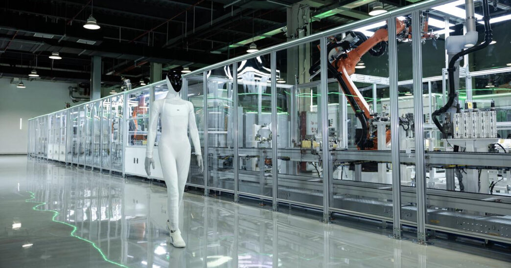 White XPENG IRON humanoid robot with a black face walking on a polished floor beside a glass-walled automated production line with orange industrial robot arms