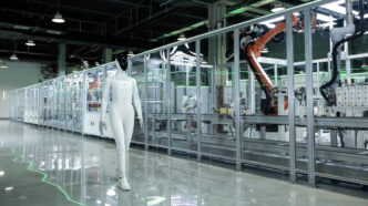 White XPENG IRON humanoid robot with a black face walking on a polished floor beside a glass-walled automated production line with orange industrial robot arms