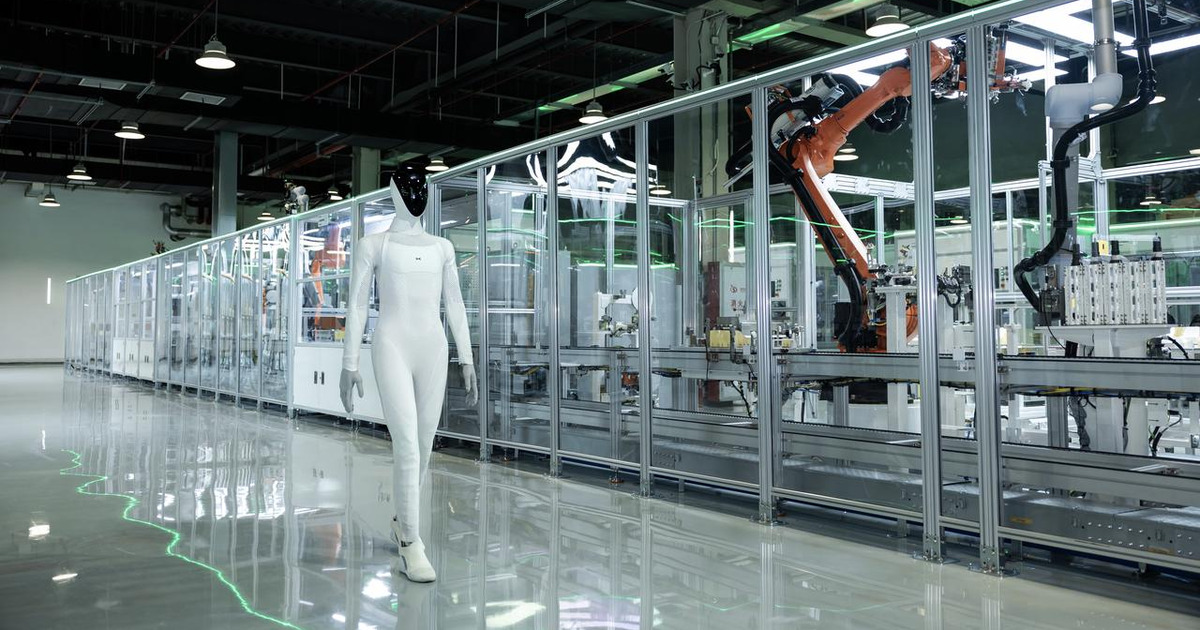 White XPENG IRON humanoid robot with a black face walking on a polished floor beside a glass-walled automated production line with orange industrial robot arms