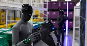 Dark grey Figure 03 humanoid robot holding a black plastic car body part beside green parts bins at BMW Group Plant Spartanburg, with a second Figure 03 working in the background