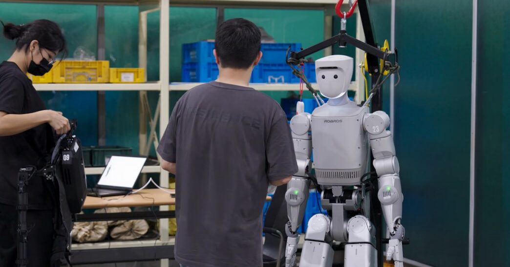 Robros IGRIS-C humanoid robot hanging from an overhead safety gantry while engineers work beside it, one wearing a teleoperation rig