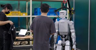 Robros IGRIS-C humanoid robot hanging from an overhead safety gantry while engineers work beside it, one wearing a teleoperation rig