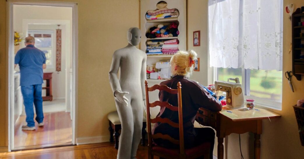 1X NEO humanoid robot in a grey knit suit standing in a home sewing room while a woman works at a sewing machine and a man walks through a doorway