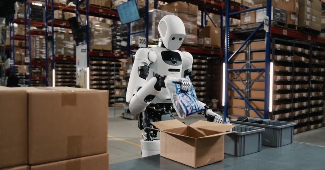 Apptronik Apollo humanoid robot packing an item into a cardboard box in a warehouse with tall shelving