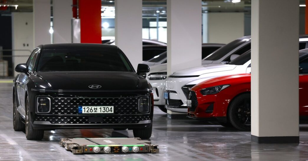 A flat Hyundai WIA parking robot with green lights on the floor in front of a black Hyundai sedan in an underground apartment car park, with white and red cars parked beside concrete pillars
