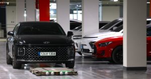 A flat Hyundai WIA parking robot with green lights on the floor in front of a black Hyundai sedan in an underground apartment car park, with white and red cars parked beside concrete pillars