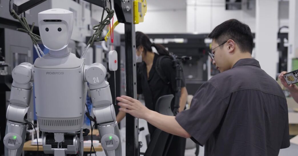 A silver Robros IGRIS-C humanoid robot with a rounded white head and the number 24 on its shoulder hangs in a support frame beside machine tools while an engineer adjusts it