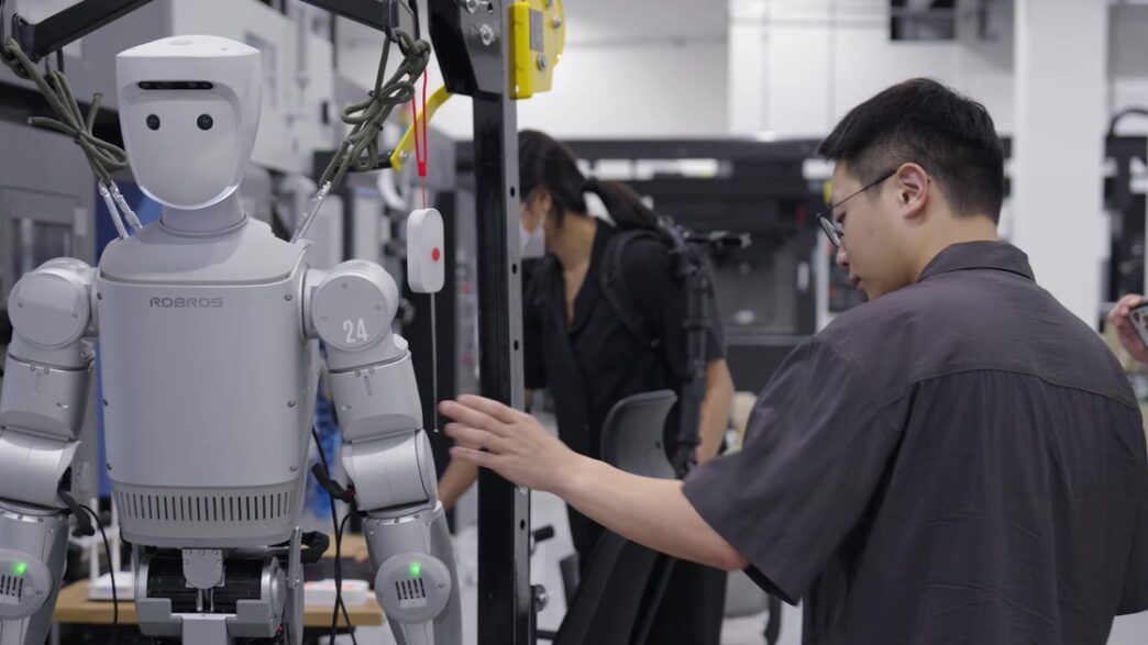 A silver Robros IGRIS-C humanoid robot with a rounded white head and the number 24 on its shoulder hangs in a support frame beside machine tools while an engineer adjusts it