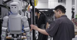A silver Robros IGRIS-C humanoid robot with a rounded white head and the number 24 on its shoulder hangs in a support frame beside machine tools while an engineer adjusts it