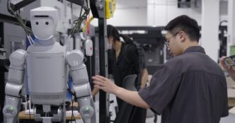 A silver Robros IGRIS-C humanoid robot with a rounded white head and the number 24 on its shoulder hangs in a support frame beside machine tools while an engineer adjusts it