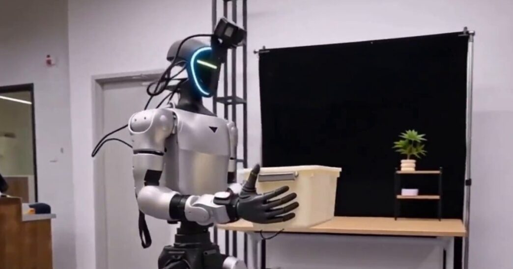 Silver Unitree G1 humanoid with a glowing visor and a head-mounted camera carrying a storage box toward a table in the Lambda-0 project video