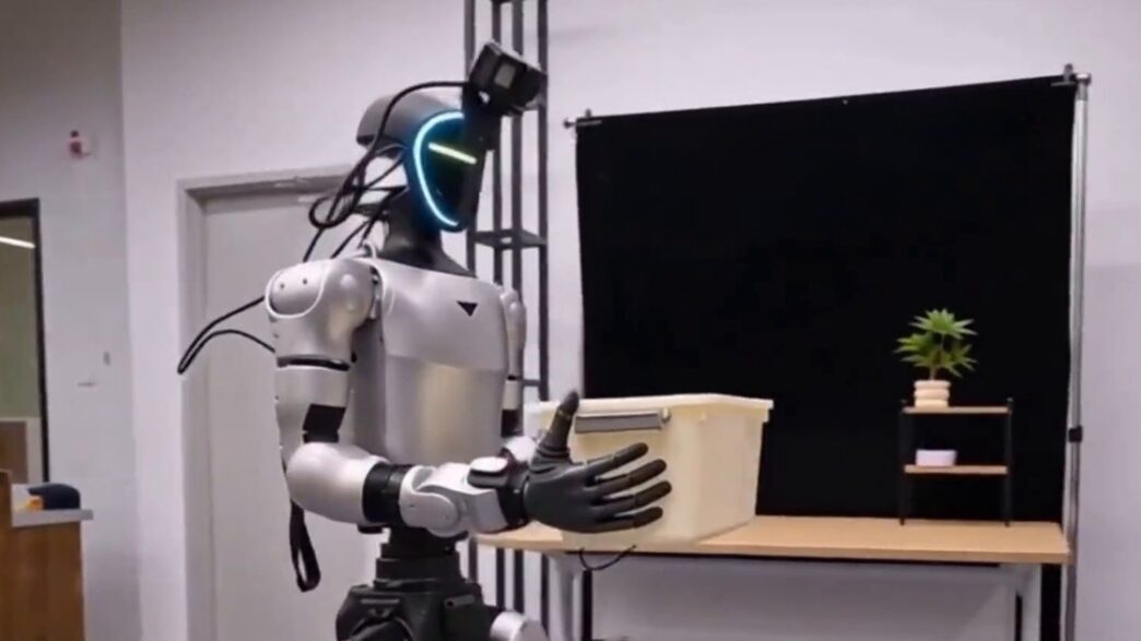 Silver Unitree G1 humanoid with a glowing visor and a head-mounted camera carrying a storage box toward a table in the Lambda-0 project video