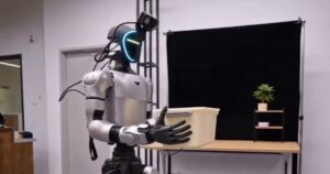 Silver Unitree G1 humanoid with a glowing visor and a head-mounted camera carrying a storage box toward a table in the Lambda-0 project video