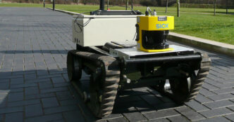 Tracked mobile robot with a yellow SICK laser scanner mounted on top, standing on a paved path next to grass