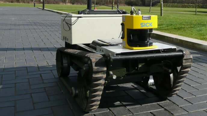 Tracked mobile robot with a yellow SICK laser scanner mounted on top, standing on a paved path next to grass