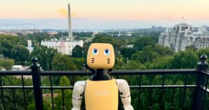 A small cream and yellow Lightberry Lumi humanoid robot with round blue eyes standing at a black railing on a rooftop in Washington, DC, with the Washington Monument and the White House grounds behind it at sunset