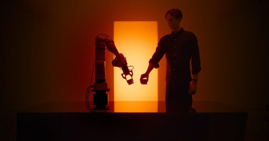 A man holding a small dark block next to a single-arm robot with a two-finger gripper, both silhouetted in front of a tall glowing orange light panel on a dark tabletop
