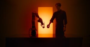 A man holding a small dark block next to a single-arm robot with a two-finger gripper, both silhouetted in front of a tall glowing orange light panel on a dark tabletop