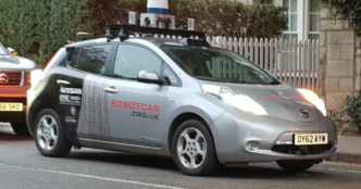 Silver Nissan Leaf research vehicle from the Oxford Robotics Institute with a roof rack of lidar units and cameras, parked on a street