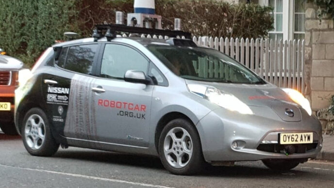 Silver Nissan Leaf research vehicle from the Oxford Robotics Institute with a roof rack of lidar units and cameras, parked on a street