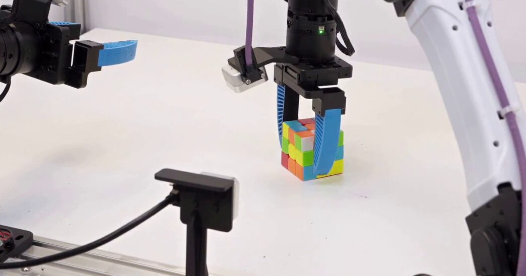 A black robot gripper with blue soft fingers grasping a Rubik's Cube on a white table, with a second robot arm and gripper reaching in from the left and a white arm on the right