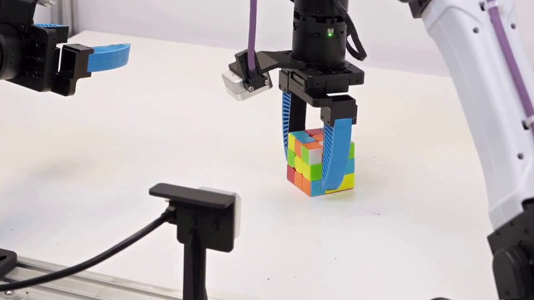 A black robot gripper with blue soft fingers grasping a Rubik's Cube on a white table, with a second robot arm and gripper reaching in from the left and a white arm on the right