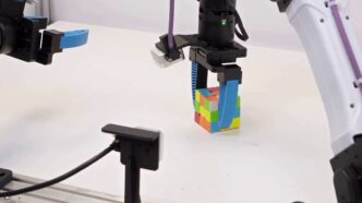 A black robot gripper with blue soft fingers grasping a Rubik's Cube on a white table, with a second robot arm and gripper reaching in from the left and a white arm on the right