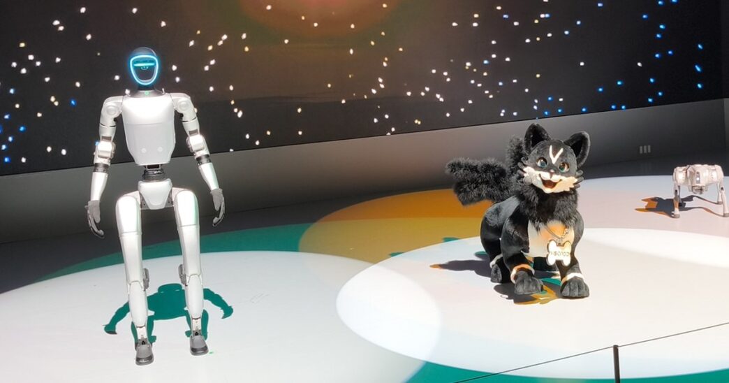 Unitree G1 humanoid robot standing on a lit stage at Japan Mobility Show 2025 beside VISIONOID's black-and-white FOX companion robot