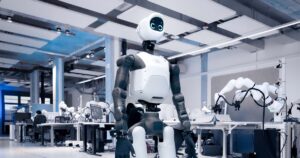 Agile Robots' white and dark gray Agile ONE SN humanoid robot with a round screen face, standing in a large robotics lab with robot arms on workbenches behind it