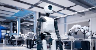 Agile Robots' white and dark gray Agile ONE SN humanoid robot with a round screen face, standing in a large robotics lab with robot arms on workbenches behind it