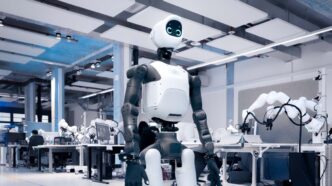 Agile Robots' white and dark gray Agile ONE SN humanoid robot with a round screen face, standing in a large robotics lab with robot arms on workbenches behind it