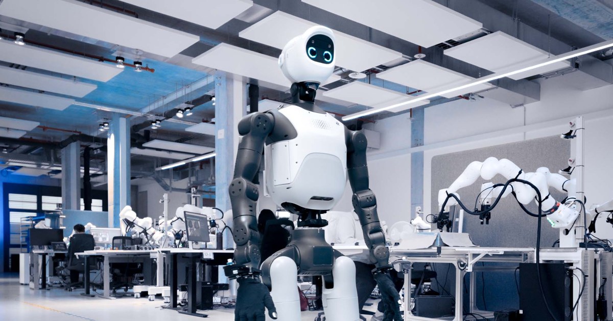 Agile Robots' white and dark gray Agile ONE SN humanoid robot with a round screen face, standing in a large robotics lab with robot arms on workbenches behind it