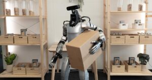 A silver humanoid robot with a black head module holding a cardboard box against its chest in front of wooden shelves